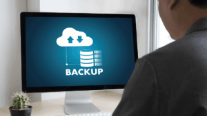 Man at a desk viewing a monitor displaying a cloud backup icon and the word BACKUP